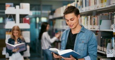 Student Of University Reading Book In Library. Man With Textbook Near Bookshelves And Students On Background. High Resolution