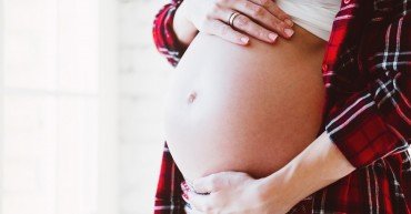 Happy beautiful pregnant woman standing at the window in bright white room and touching her belly.