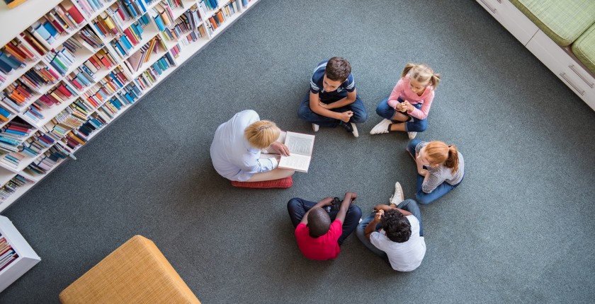 Teacher reading fairy tales to children sitting in a circle at library. Top view of librarian sitting with five multiethnic children on floor. Teacher reading book to cute girls and young boys at school.