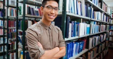 Portrait of a smiling asian man standing with arms folded in university library and looking at camera